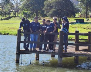 Em um deck de madeira às margens de um lago, servidores do Imasul participam de treinamento prático para uso de uma nova sonda multiparâmetro. O grupo segura equipamentos ligados por cabos, com sensores submersos na água para medição de parâmetros ambientais. Os participantes usam camisetas azuis da instituição. Ao fundo, há árvores, gramado e um prédio alto sob céu azul e ensolarado.