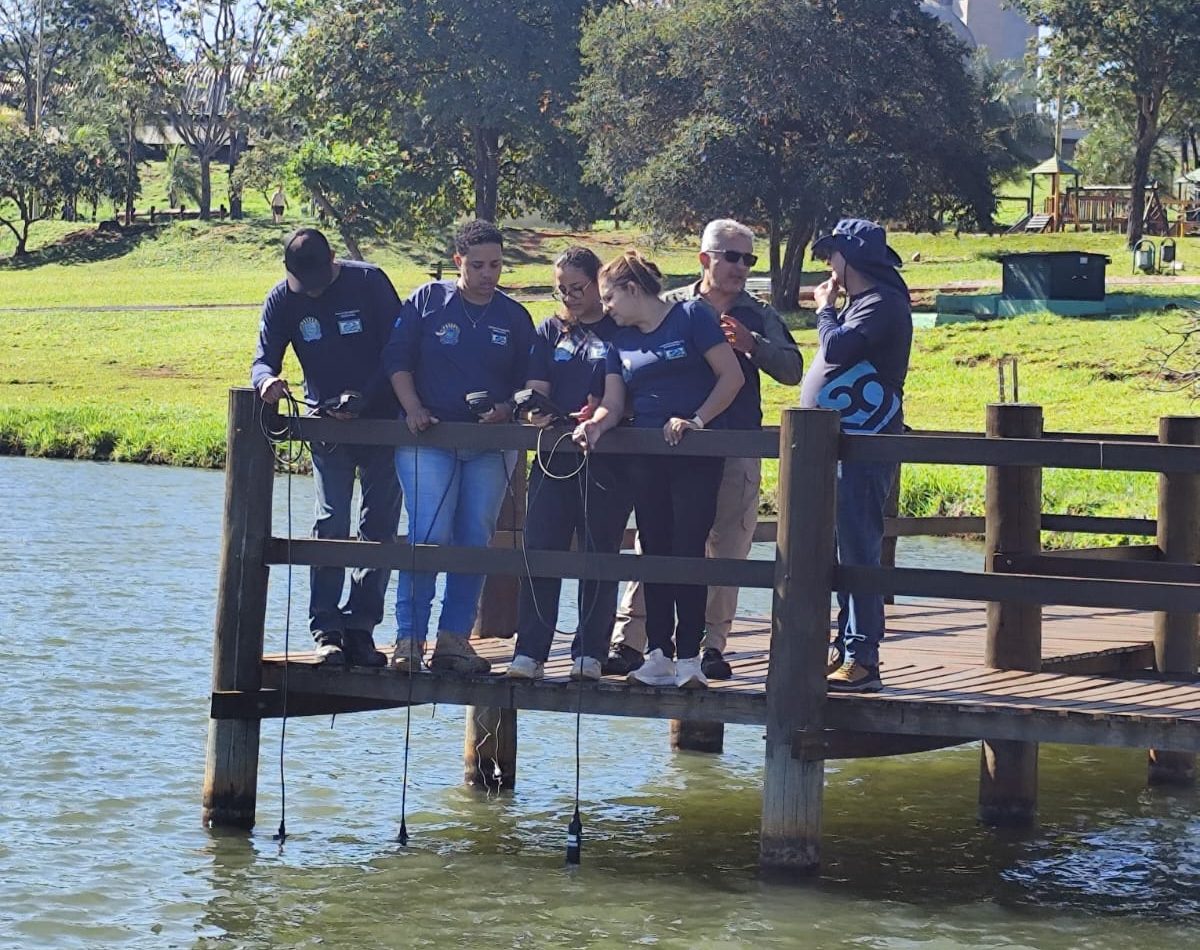 Em um deck de madeira às margens de um lago, servidores do Imasul participam de treinamento prático para uso de uma nova sonda multiparâmetro. O grupo segura equipamentos ligados por cabos, com sensores submersos na água para medição de parâmetros ambientais. Os participantes usam camisetas azuis da instituição. Ao fundo, há árvores, gramado e um prédio alto sob céu azul e ensolarado.